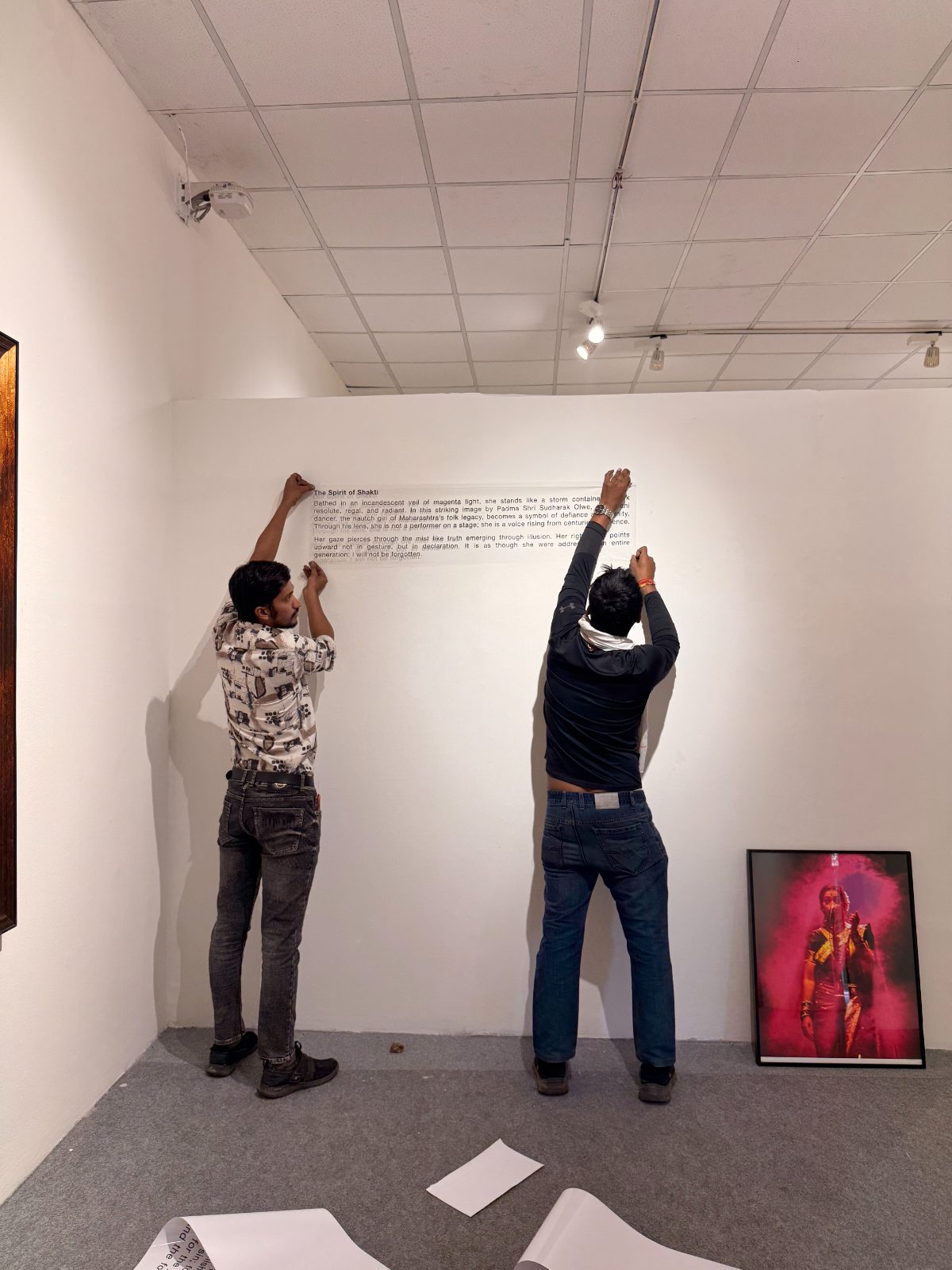Two people fixing The Spirit of Shakti wall text, the magenta portrait leaning below